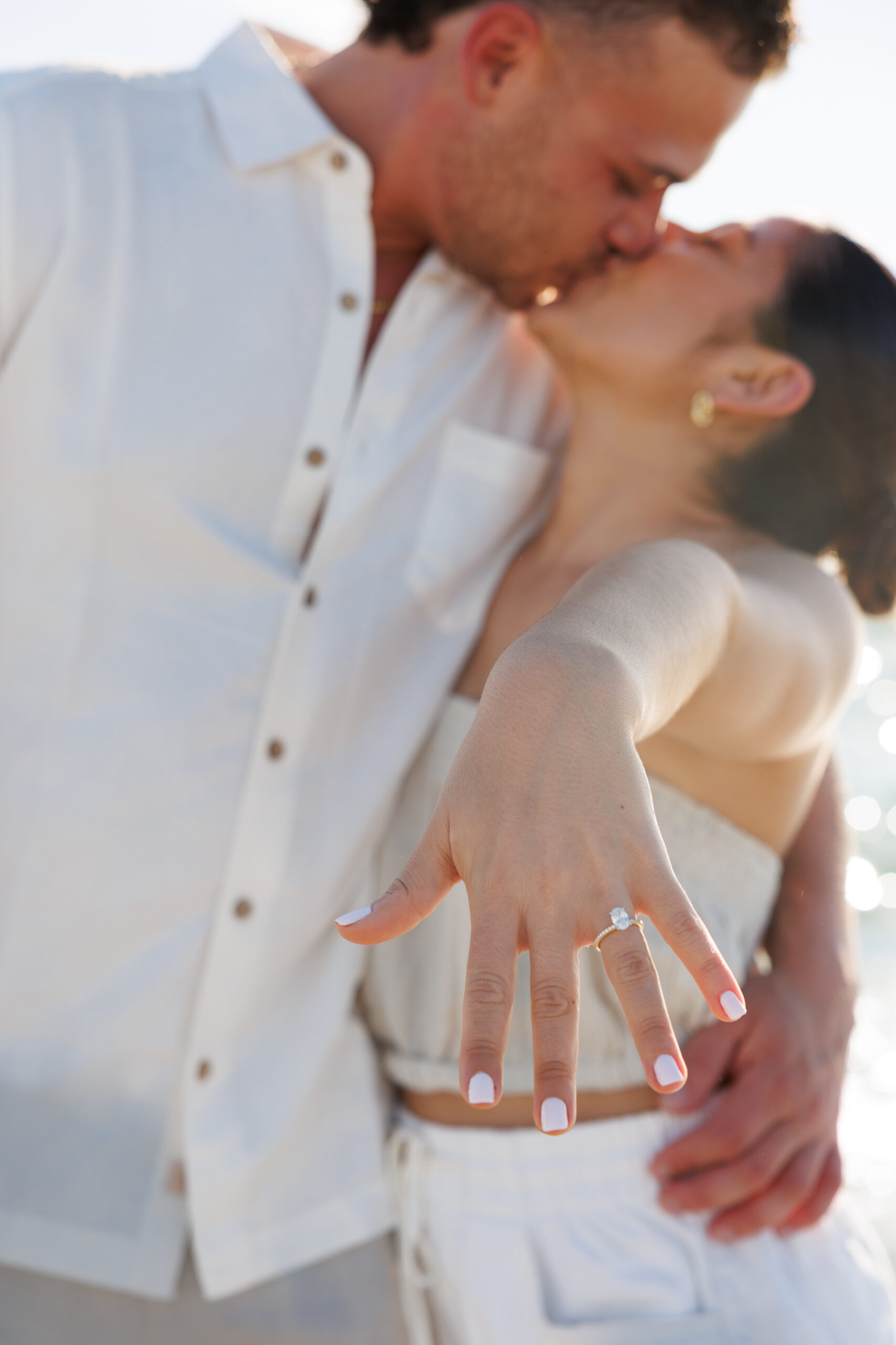 7N4A0374 A pregnant woman in a white lace dress cradles her baby bump while holding a vibrant sunflower surrounded by greenery.