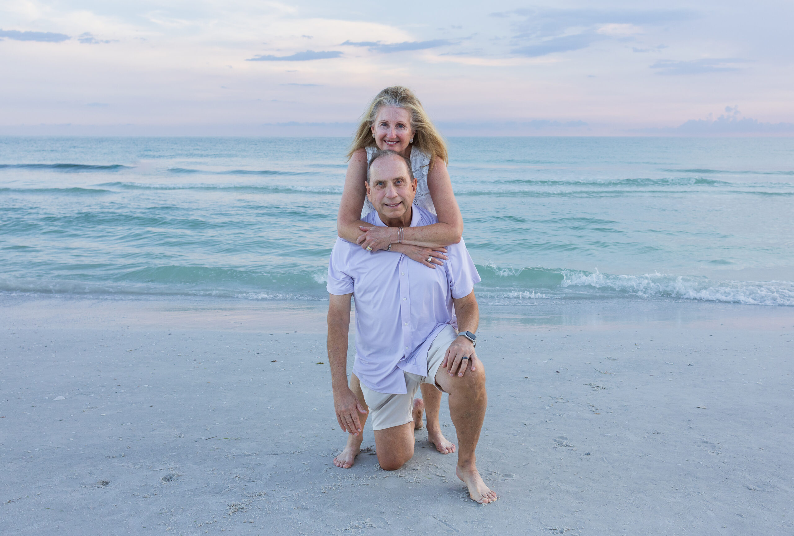 7N4A5702 A pregnant woman in a white lace dress cradles her baby bump while holding a vibrant sunflower surrounded by greenery.